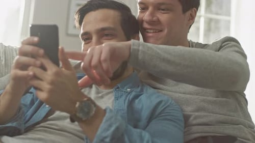 Cute Male Gay Couple Spend Time at Home. They are Lying Down on a Sofa and Use a Smartphone. They B
