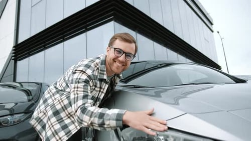 Happy young man buying a car in showroom outdoors, guy hugging hood of new car