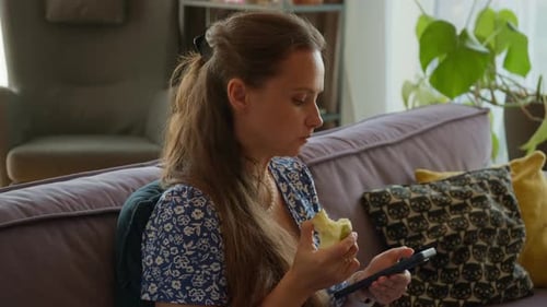 Woman Eating Apple Using Tablet on Couch Indoors