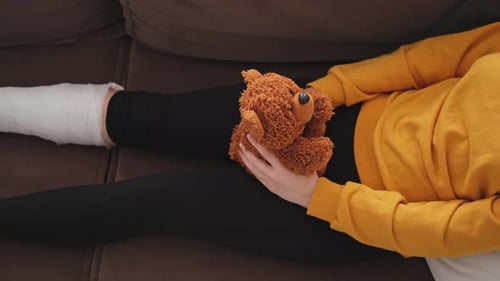 Junges Mädchen mit gebrochenem Beinabdruck sitzt auf dem Sofa Trauriges kleines Kind umarmt ihren Teddybär und erholt sich