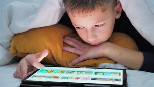 Little Boy Plays on Tablet While Lying Under Blanket