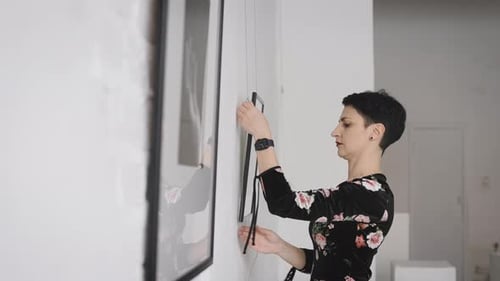 A Woman Gallerist Hangs New Paintings and Collages on the Wall of the Exhibition Hall