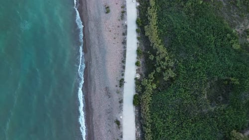 Zenithal view of a beach with waves hitting the shore at sunset, as people walk and exercise along t