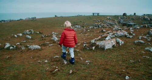 Child Walk on Ocean Shore Meadow on Rainy Day on Vacation Trip in Orange Jacket