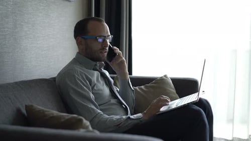 Man Using Laptop and Talking on Phone on Couch