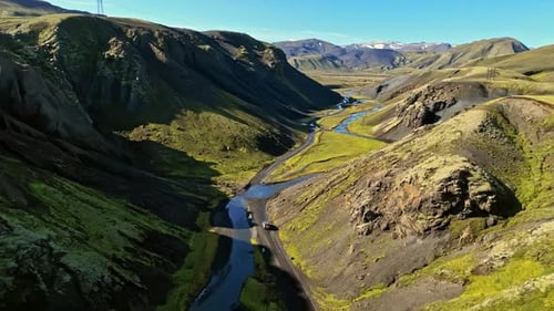 Drone view of vehicles following a narrow road through a steep mossy canyon with a winding river,