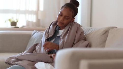 Jeune femme afro-américaine malade bouleversée enveloppée dans une couverture prenant des mouchoirs et se mouchant librement