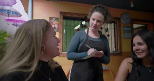 Restaurant server standing beside table writing order while friends wait and chat showing everyday