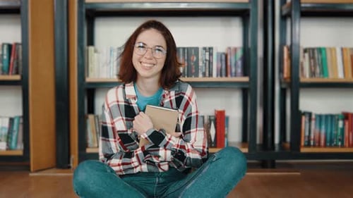 Portrait of Young Woman Student Reading a Book in the Library Sitting on the Floor Students in a