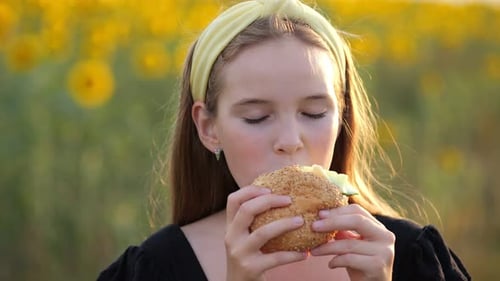 Teen Girl Eats Hamburger Against Blurry Sunflower Field