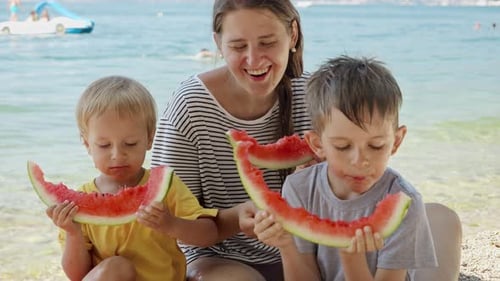 Slow-motion of a joyful mother and her two boys eating sweet watermelon by the sea