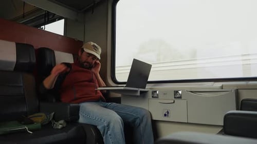 Ein Mann sitzt am Zugfenster, konzentriert auf seinen Laptop, während draußen die verschwommene Landschaft vorbeirauscht