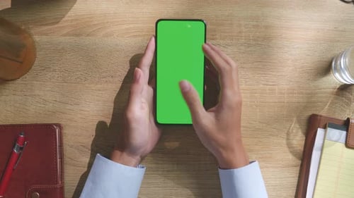 Top down view of man's hands using green screen smartphone on a wooden desk