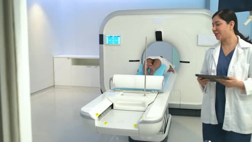 Technologist tapping tablet is initiating CT scan, patient is sliding into gantry for brain imaging