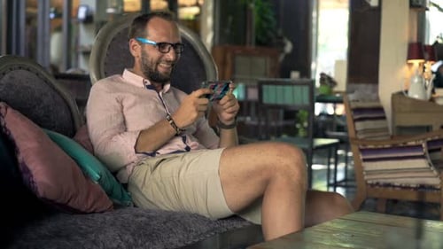 Happy Man Plying Game on Smartphone Sitting on Sofa in Trendy Cafe 30s