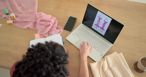 Seeing laptop sketches woman designer smoothing pink fabric at table and noting design in notebook