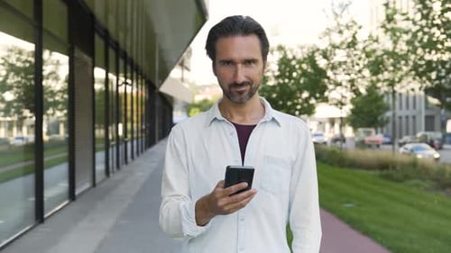 A Caucasian Man Looks at a Smartphone and Then Smiles at the Camera in a Street in an Urban Area