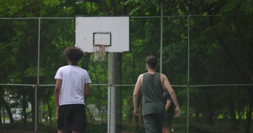 Group of young multiethnic men playing basketball on outdoor court surrounded by trees, African