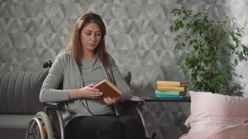 Young Woman Takes Book with Brown Cover From Stack on Table