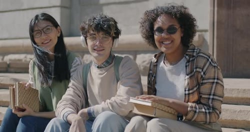 Cheerful Youth Students Sitting Outdoors in Street Holding Books Enjoying Summer Day in City Smiling