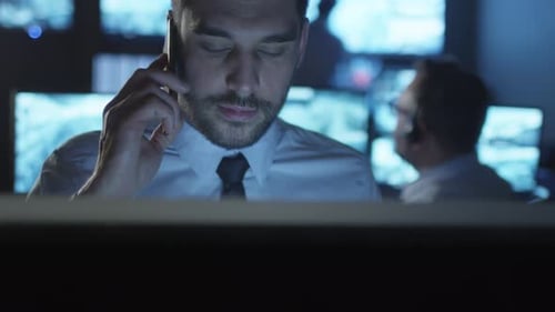 Security officer is talking on the phone while working on a computer in a dark monitoring room fill