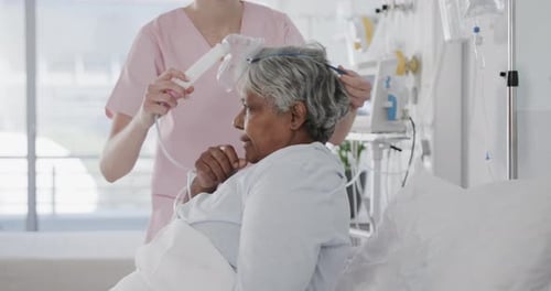 Diverse female doctor giving oxygen mask to senior female patient in hospital bed, slow motion