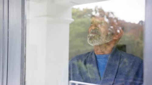 Happy senior biracial man looking through window, unaltered, in slow motion