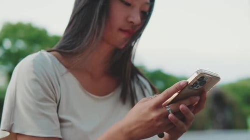 Close up, young woman sits on bench in city park, uses mobile phone
