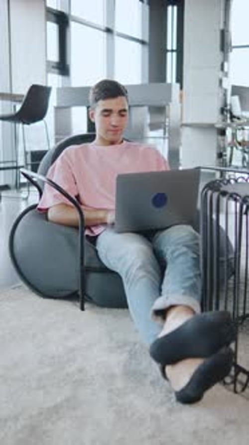 Young Man Works on Laptop While Sitting in Cozy Chair Inside Modern Workspace During Daytime
