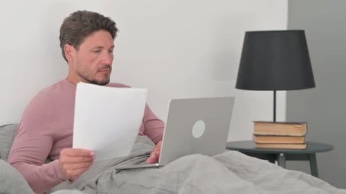 Man Working With Laptop and Paper in Bed