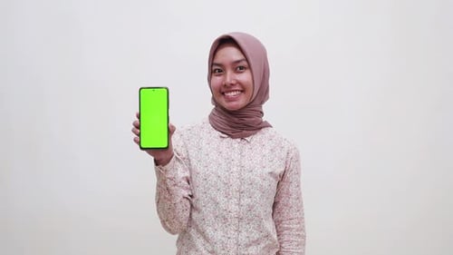 Happy young asian muslim woman standing while presenting a blank green screen cell phone display. Is