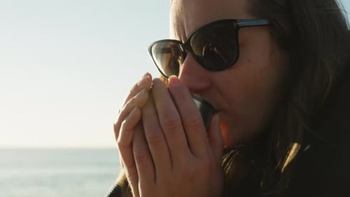 A cool Morning at the Sea, a young woman warms her hands holding a hot mug with a drink, steam is co