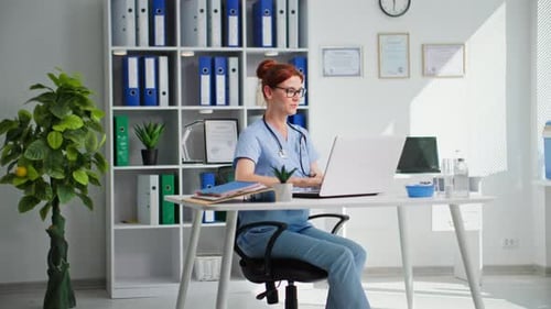 Young Beautiful Female Doctor in Glasses Works at a Laptop in a Medical Office Smiles and Looks at