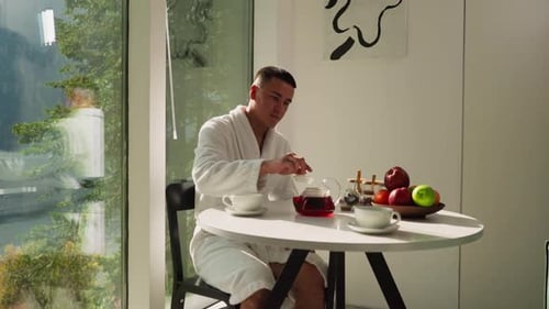 Un homme met une casquette sur une théière assis dans la cuisine