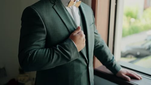 Man in a green suit adjusting his lapel while standing by a window.