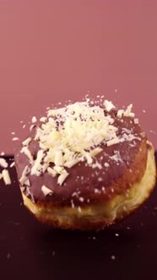 Vertical slow-motion shot of a Berliner donut falling, with white shavings scattering dramatically.