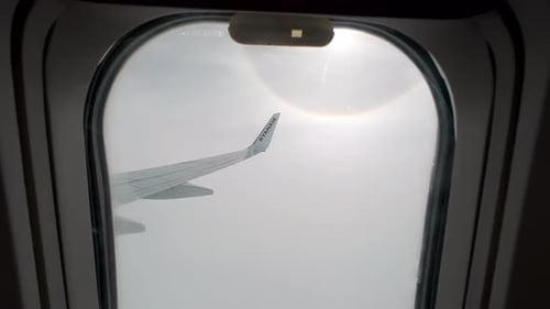 Smooth push in between airplane window to view of wing tip against clouds, sunburst around sun