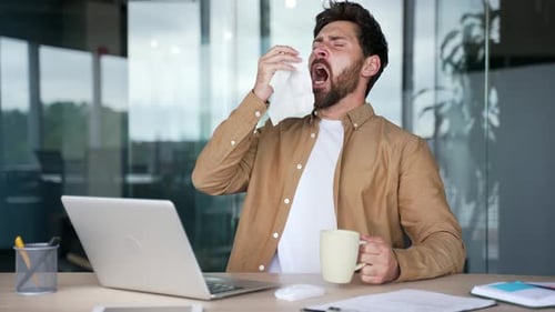 Sick businessman suffers from a cold while sitting at a desk at a workplace in a business office.