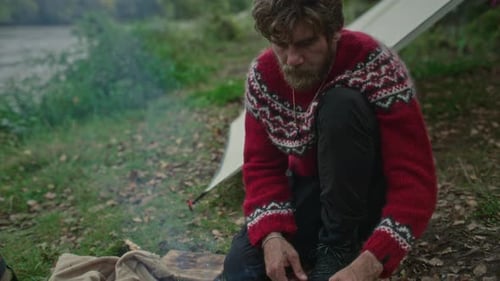 Man Roasting Skewered Meat over Fire at Forest Campsite by River