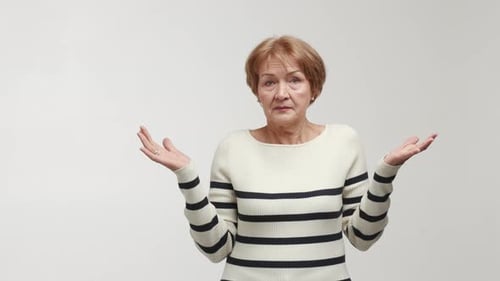 Uma mulher idosa com um olhar confuso encolhe os ombros balança a cabeça levanta as sobrancelhas, abre os braços e as palmas das mãos para