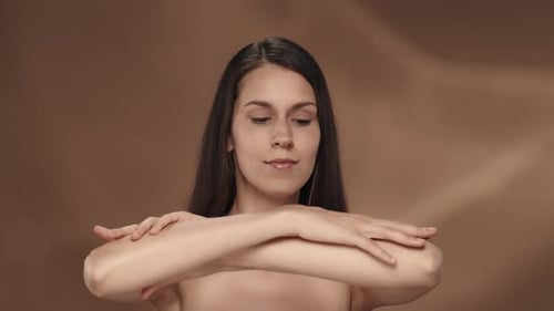 A Seminude Woman Enjoys the Velvety and Softness of the Skin of Her Hands in the Studio on a Brown
