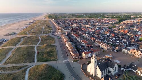 Aerial view of a seaside town at sunset, showing red-roofed houses, a historic church, sandy dunes
