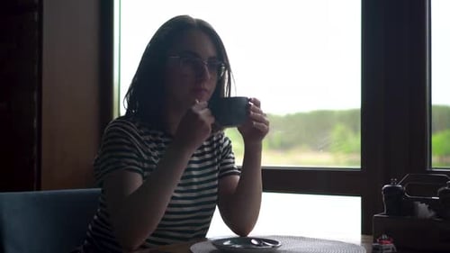 A Young Woman Sits in a Cafe By the Window and Drinks Coffee Panoramic Window Overlooking the River