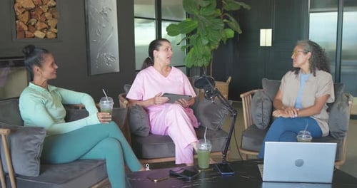 Diverse women hosting in lounge, right host leading, center reading tablet, microphone present