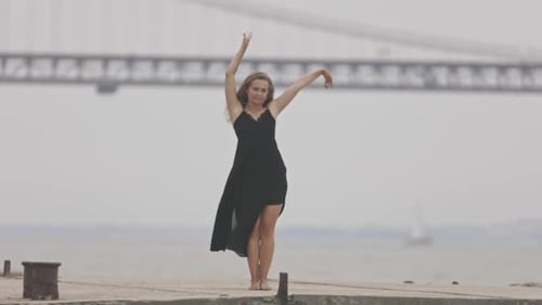 An Adult Carefree Woman in Black Dress Dancing on the Pier in Slow Motion with a Bridge on the