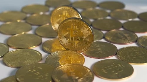 Zwei goldene Bitcoin-Münzen stehen zwischen vielen liegenden Münzen, die sich kreisförmig auf weißem Grund drehen.