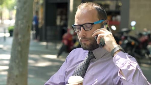 Young Businessman with Coffee Talking on Cellphone Sitting on Bench in City 30s