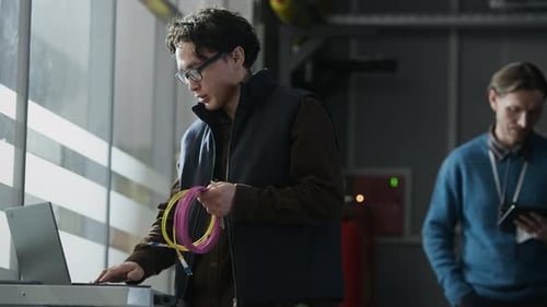 Asian Network Engineer Holding Cable Coil while Working on Laptop in Data Center