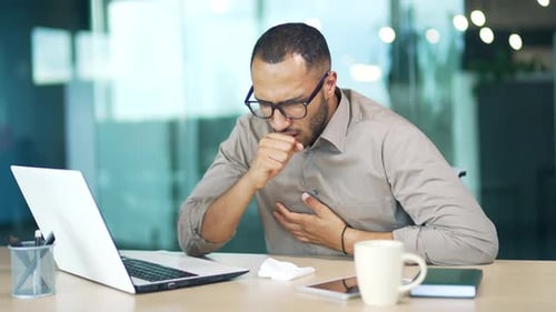 Young adult man coughing while sitting at workplace at desk in modern office. Sick with a cold