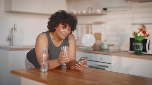 Woman Drinking Water While Using Phone in Kitchen